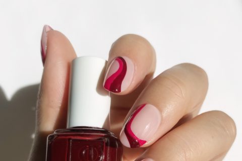 a hand with abstract nail polish designs holding a berry coloured bottle of Essie nail polish